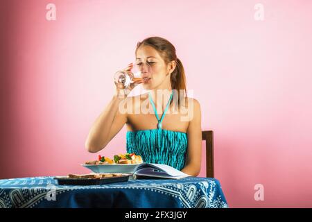 Attraktive junge alleinstehende Frau sitzt am Tisch mit Essen und offenem Buch und trinkt Wein aus Glas. Auf pastellrosa Hintergrund. Stockfoto