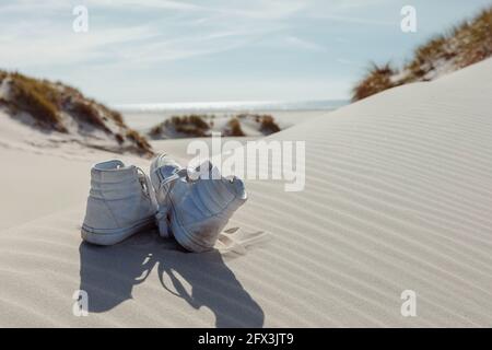 Ein paar weiße Sneakers am Strand auf Sand gelassen. Dünen mit grünem Gras Vegetation. Sommerurlaub, Urlaub am Meer Konzept. Stockfoto