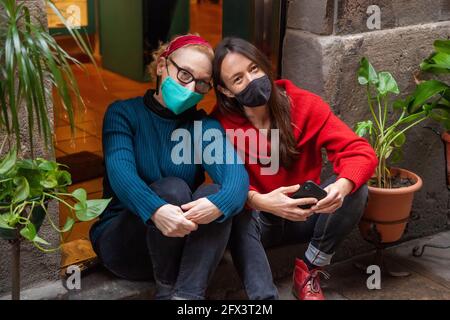 Porträt zweier Freundinnen mittleren Alters mit schützenden Gesichtsmasken, die dicht beieinander am Eingang ihres Ladens sitzen. Konzept: Arbeiten während der cor Stockfoto