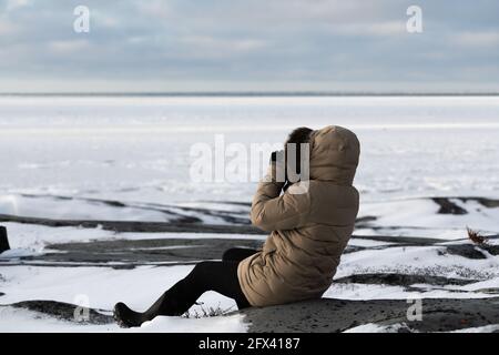 Frau, die während der Eisbärsaison auf Eisfelsen am Rande der Hudson Bay in Churchill, Manitoba, Kanada, sitzt und die Tundra-Meereislandschaft fotografiert Stockfoto