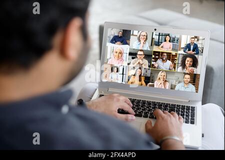 Junger Geschäftsmann mit Laptop-App für Video verbinden Sie sich mit Kollegen, nimmt an Videokonferenzen, Bildschirm mit vielen verschiedenen Menschen in Video-Meeting beteiligt, Gruppe von multirassischen Kollegen auf dem Bildschirm Stockfoto