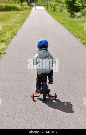 Rückansicht eines Kindes mit blauem Helm, das auf einer Straße ein Fahrrad mit Trainingsrädern fährt. Stockfoto