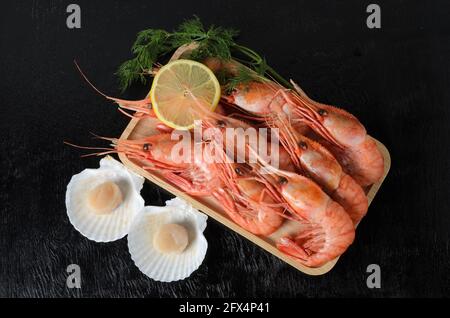 Gekochte Garnelen in einer Schale mit Dill und Zitrone in einer Holzschüssel mit einer rohen Jakobsmuschel in einer Schale auf dunklem Holzgrund. Selektiver Fokus. Stockfoto