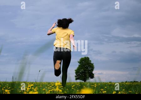 Ein Mädchen in einem gelben T-Shirt bewegt sich mit Blumen über das Feld. Gelbe Blüten, grünes Gras, blauer Himmel, Eiche. Reisen Sie außerhalb der Stadt. Freizeit. Stockfoto