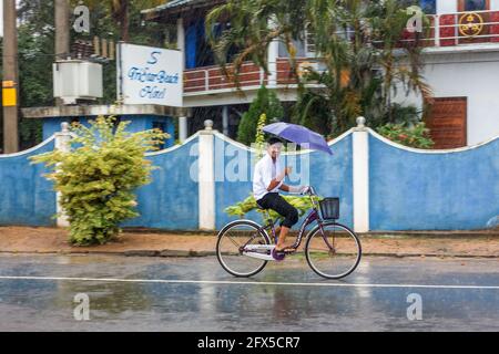 Der Sri-lankische Mann fährt mit dem Fahrrad und trägt einen Regenschirm bei sintflutartigen Regenfällen (aufgenommen mit der „Schwenk“-Kameratechnik), Arugam Bay, Eastern Province, Sri Lanka Stockfoto