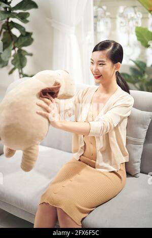 Bester Freund zum Umarmen. Schöne junge asiatische Frau umarmt ihren Teddybär mit Lächeln, während auf der Couch sitzen Stockfoto