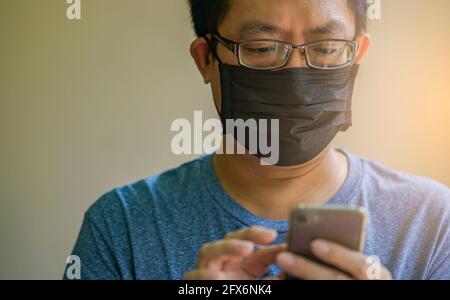 Porträt Asian Mann trägt schwarze medizinische Maske und SMS auf Smartphone, Nahaufnahme Asian Mann mit Brille, schwarze kurze Haare, Augen zu smartphon schauen Stockfoto