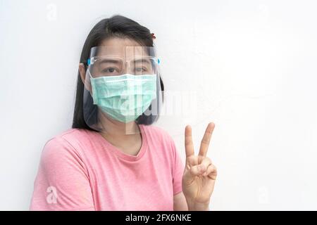 Isoliertes Porträt der asiatischen Frau trägt medizinische Gesichtsmaske und Gesichtsschutz auf weißem Hintergrund, zeigt Geste zwei Finger, schwarze kurze Haare und tragen Stockfoto