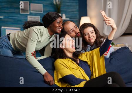 Multiethnische Freunde machen lustige Gesichter, während sie Selfie-Fotos machen, feiern in der Wohnung, trinken Bier. Eine Gruppe von verschiedenen Menschen lachend, sitzen auf dem Sofa spät in der Nacht im Wohnzimmer. Stockfoto