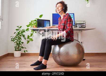 Frau mit Telearbeit, die vor ihr auf einem Fitball sitzt Schreibtisch, der über ein Headset spricht Stockfoto