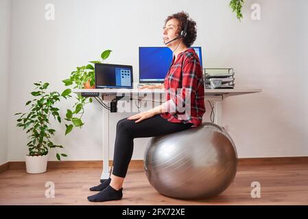 Frau mit Telearbeit, die vor ihr auf einem Fitball sitzt Schreibtisch, der über ein Headset spricht Stockfoto