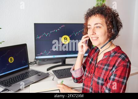 Weibliche Händler operiert mit Bitcoins von ihrem Stehtisch aus Sprechen über das Mobiltelefon und Tragen eines Headsets Stockfoto