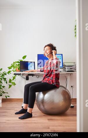 Frau mit Telearbeit, die vor ihr auf einem Fitball sitzt Schreibtisch, der über ein Headset spricht Stockfoto