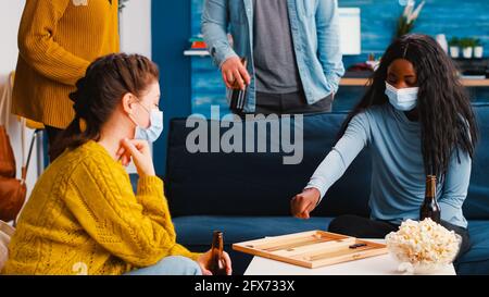 Fröhliche Frau mit Würfeln, die Backgammon mit multiethnischen Freunden spielen Gesichtsmaske als Prävention gegen Covid 19-Virus während der globalen Pandemie sitzt auf der Couch, trinkt Bier und isst Popcorn Stockfoto