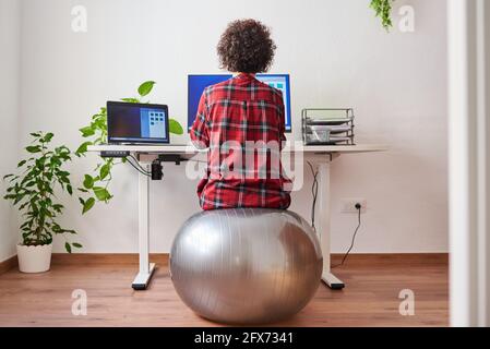 Rückansicht einer Telearbeiterin, die auf einem Fitball sitzt Vor ihrem Schreibtisch Stockfoto