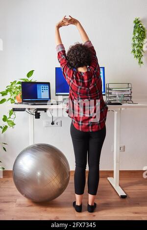 Frau streckt sich während der Arbeit an einem verstellbaren Schreibtisch neben einem Fitball Stockfoto