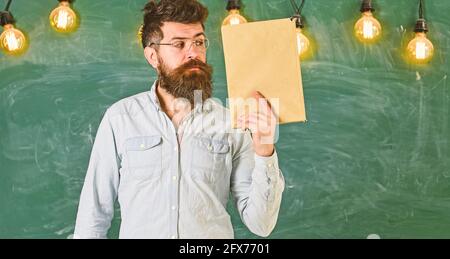 Selbstbewusster Lehrer oder Geschäftsmann, der vor der Tafel steht, hält das Buch in der Hand. Wissenschaftler in Brillen hält Buch, Tafel auf Hintergrund Stockfoto