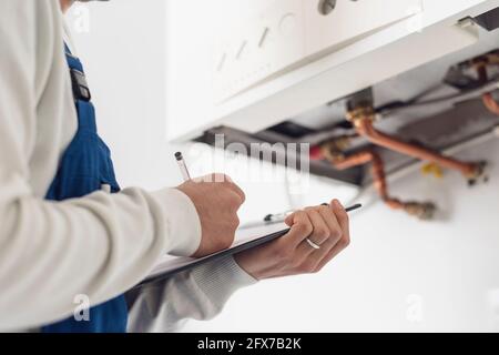 Professioneller Klempner, der zu Hause einen Kessel überprüft und schreibt Eine Zwischenablage Stockfoto
