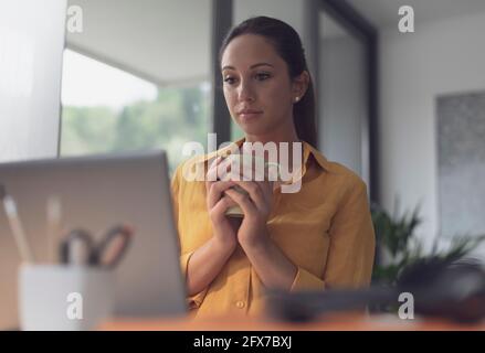 Junge Frau, die am Schreibtisch sitzt und Kaffee trinkt, starrt auf den Laptop-Bildschirm Stockfoto