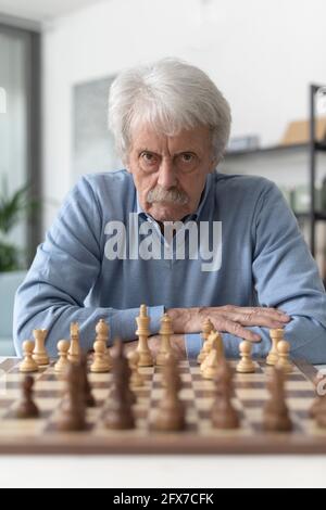 Selbstbewusster älterer Mann, der zu Hause Schach spielt, schaut auf die Kamera Stockfoto