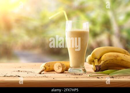 Banana Smoothie in Glas mit Stroh auf Holztisch mit Obst und Plantage im Hintergrund. Vorderansicht. Stockfoto