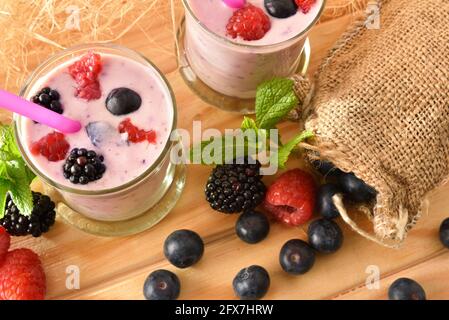 Waldbeermilchshake in Glasgläsern auf Holztisch mit Sack voller Heidelbeeren, Brombeeren Himbeeren mit grünem Waldhintergrund. Draufsicht Stockfoto