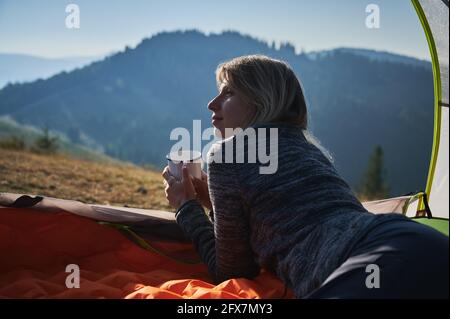 Nahaufnahme der Rückansicht eines hübschen Mädchens, das sich beim Wandern in den Bergen in ihrem Zelt entspannt, Metallbecher in den Händen hält und links friedlich zuschaut. Stockfoto