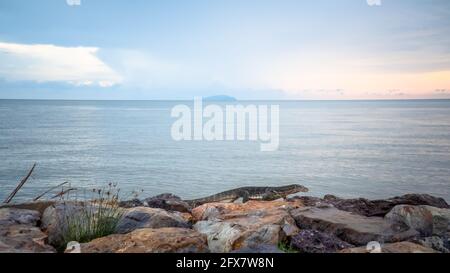 Asiatischer Wassermonitor Eidechse kriechen und gehen auf felsigen Steinen mit verschwommenem Meereshintergrund und Sonnenuntergang Himmel und Wolke, Pontian, Malaysia. Stockfoto