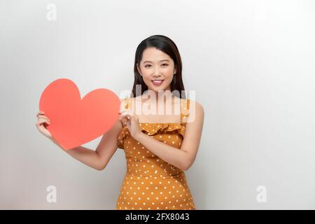 Lächelnd hübsche Frau, die nach oben schaut, großes rotes Herz an der Brust hält, Tagträumend von Männern in der Liebe, Stockfoto