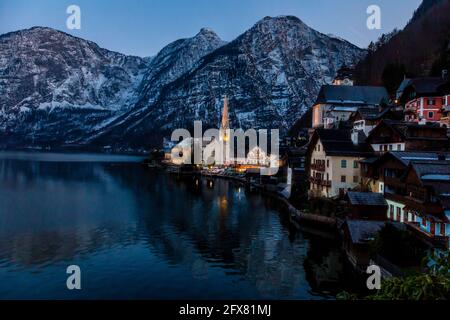 Blick auf den schönen Hallstätter See und die berühmte Kirche in letzter Zeit Abend nach Sonnenuntergang im frühen Frühling mit bedeckten Bergketten In etwas Schnee im Hintergrund Stockfoto