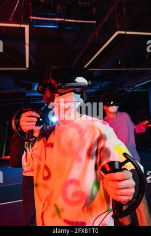 Teenager in medizinischen Maske Gaming in der Nähe verschwommen Freunde in vr-Wiedergabezone Stockfoto