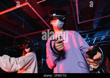 Teenager in medizinischer Maske und vr Headset-Gaming in der Nähe Freund in der Spielzone Stockfoto