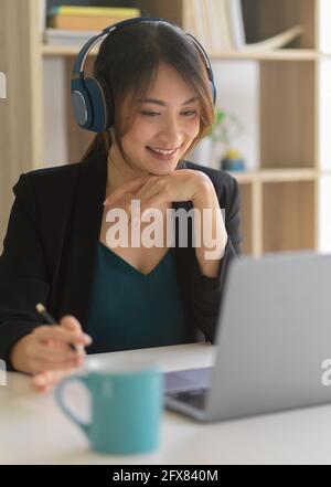 Kurze Aufnahme einer glücklichen Geschäftsfrau, die lächelt und Kopfhörer verwendet Arbeiten mit einem Laptop im Büroraum Stockfoto