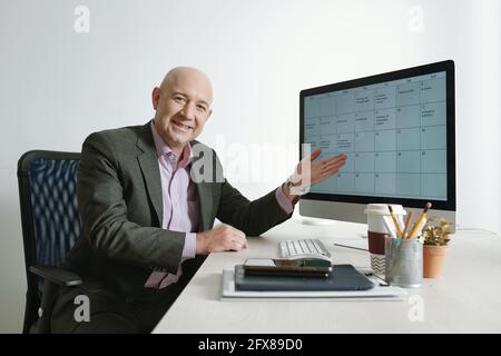 Porträt eines fröhlichen, kahlköpfigen und reifen Geschäftsmannes im Anzug Auf dem Computermonitor auf seinen Zeitplan zeigen Stockfoto
