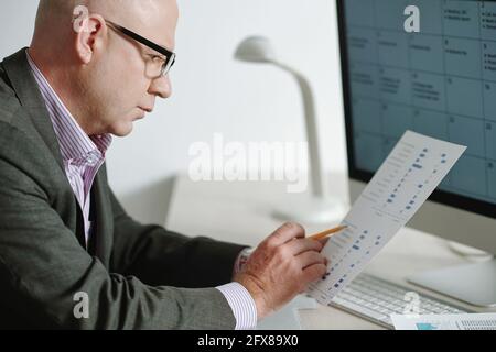 Konzentrierter reifer Geschäftsmann in Brillen, der am Schreibtisch mit Computer sitzt Und Statistiken auf Papier anzeigen Stockfoto