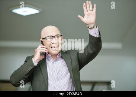 Aufgeregt kahlköpfigen kaukasischen Geschäftsmann in Brillen und formellen Anzug reden Per Telefon und mit der Hand winken, während der Mitarbeiter begrüßt wird Stockfoto