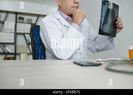 Nahaufnahme eines nachdenklichen Radiologen in weißem Mantel, der am Tisch sitzt Und die Analyse von Röntgenbildern im Büro, während Sie über Diagnose nachdenken Stockfoto