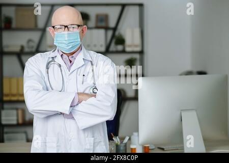 Portrait von selbstbewussten erfahrenen reifen kaukasischen Arzt in Brillen und Maske steht mit gekreuzten Armen im modernen Büro Stockfoto