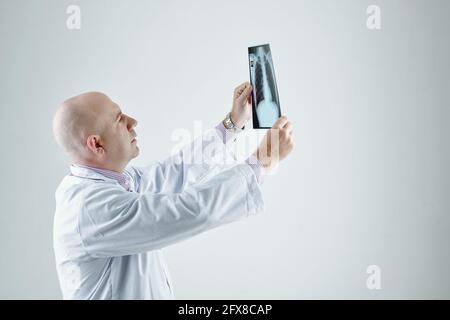 Konzentrierter Weißkopfradiologe in weißem Fell, der vor grauem Hintergrund steht Und die Anzeige des Röntgenbildes gegen Licht Stockfoto