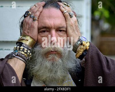 Der alte esoterische kaukasische Weise Mann mit dickem grauem Bart posiert für die Kamera, hält seinen Kopf in den Händen und zeigt seine sortierten Ringe und Armbänder. Stockfoto