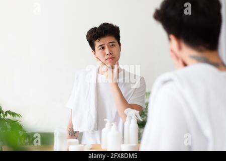Fröhlicher asiatischer junger Mann mit Tätowierungen in weißem T-Shirt, mit Handtuch, berührt Hand an Wange, karrt Stoppeln Stockfoto