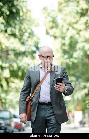 Serious beschäftigt reifen kahlen Geschäftsmann mit Ledertasche mit Smartphone Während der Arbeit auf Bewegung im Sommer Stockfoto