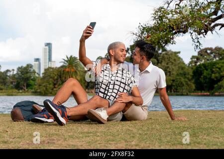 Junge lateinamerikanische schwule Paare, die Selfies machen, sitzen auf dem Gras am See in einem öffentlichen Park. Stockfoto