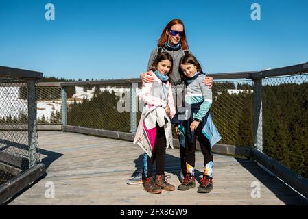 Porträt einer Familie, die im Winter den Weg zur Aussichtsplattform mit Holzkäfeln besucht. Familie mit Kindern über Baumkronen auf Holzbalkon Steg durch die Stockfoto