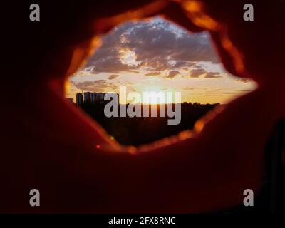 Blick durch die Hände in Form eines imaginären Spiglases über den atemberaubenden orangefarbenen Sonnenuntergang über der Stadt mit Gebäudesilhouetten. Nächtliches industrielles Stadtbild. Stockfoto