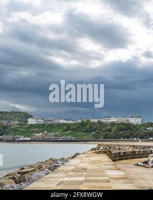 Eine Seeszene mit einem Hügel und weißen Gebäuden auf der Skyline. Ein Pier und Wellenbrecher sind im Vordergrund und dunkle Wolken sind oben. Stockfoto