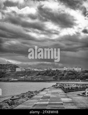 Eine Seeszene mit einem Hügel und weißen Gebäuden auf der Skyline. Ein Pier und Wellenbrecher sind im Vordergrund und dunkle Wolken sind oben. Stockfoto