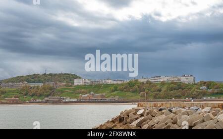 Eine Seeszene mit einem Hügel und weißen Gebäuden auf der Skyline. Ein Pier und Wellenbrecher sind im Vordergrund und dunkle Wolken sind oben. Stockfoto