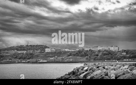 Eine Seeszene mit einem Hügel und weißen Gebäuden auf der Skyline. Ein Pier und Wellenbrecher sind im Vordergrund und dunkle Wolken sind oben. Stockfoto