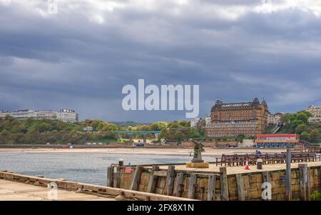 Eine Seeszene mit einem Hügel und Gebäuden auf der Skyline. Ein Pier mit einem alten Gewehr ist im Vordergrund und ein Strand ist dahinter. Ein dunkler Himmel ist oben. Stockfoto